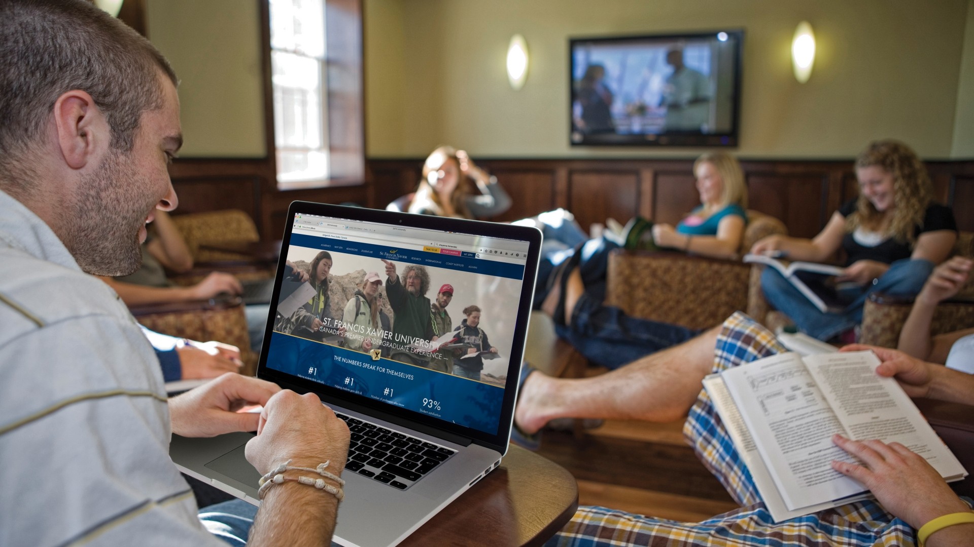 Students studying in the Bishops Hall lounge area. They are sitting on chairs with a small table in the center and a TV on the wall.
