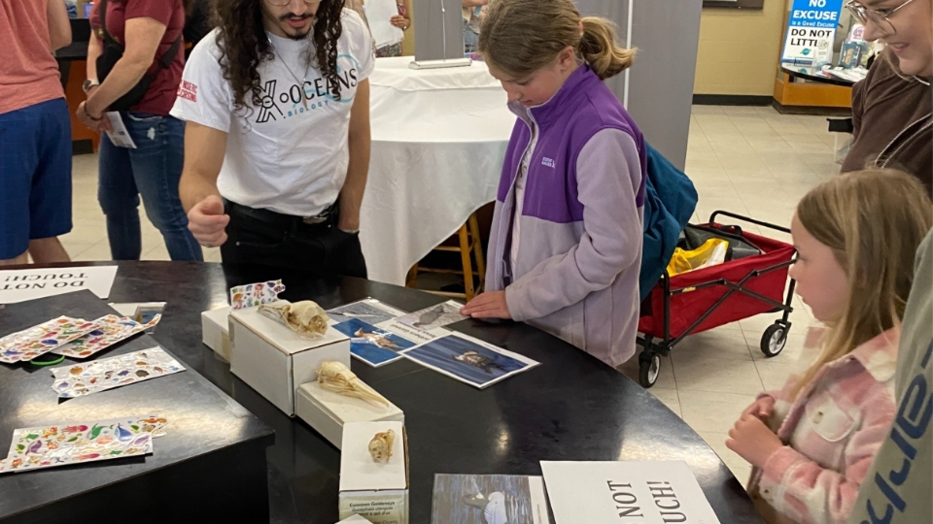People gathered around a display table with bird photos and labeled boxes, guided by a 'ZOO TEEN' presenter.