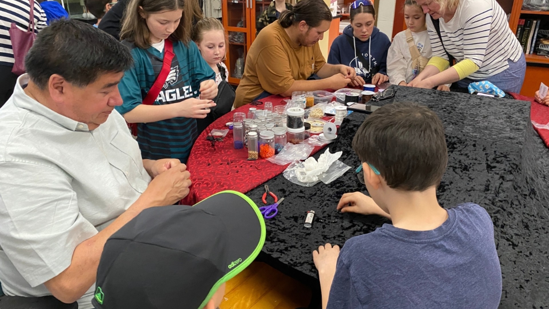 Children and adults participating in a beadwork activity at a table covered with crafting supplies, in an Indigenous education setting.