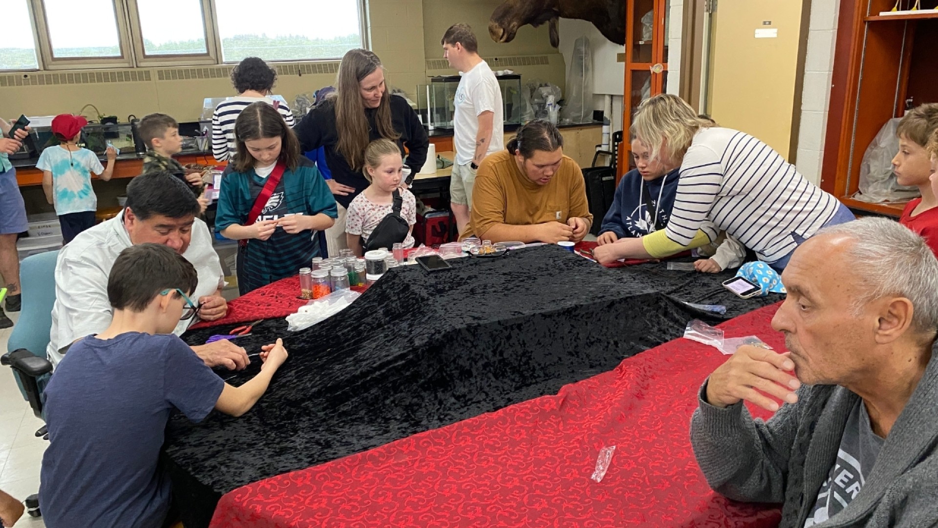People gathered around a table with red and black cloths, participating in an activity related to Indigenous education.