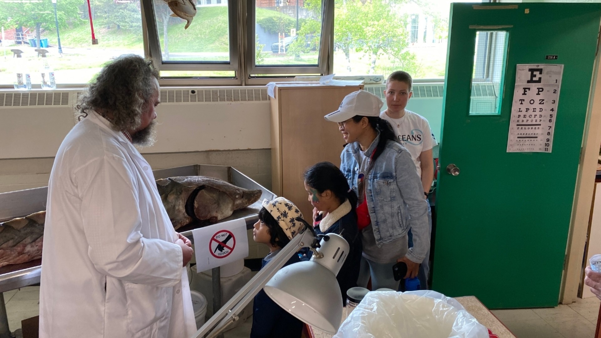 A man in a lab coat presenting a large fish to a group in a science classroom with lab equipment and an anatomical skeleton.