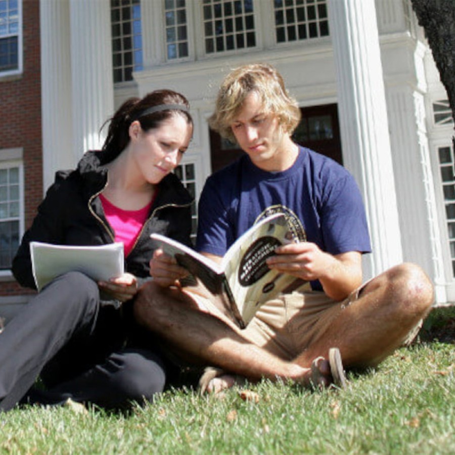 Two business students sitting on the lawn outside of a residence building reviewing their textbook