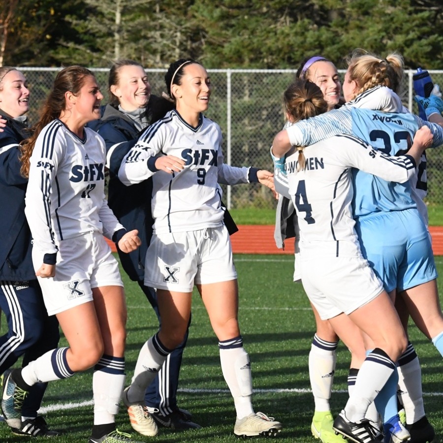 StFX X-Womens soccer players in white long sleeve uniforms running to celebrate with their goalie in a blue uniform