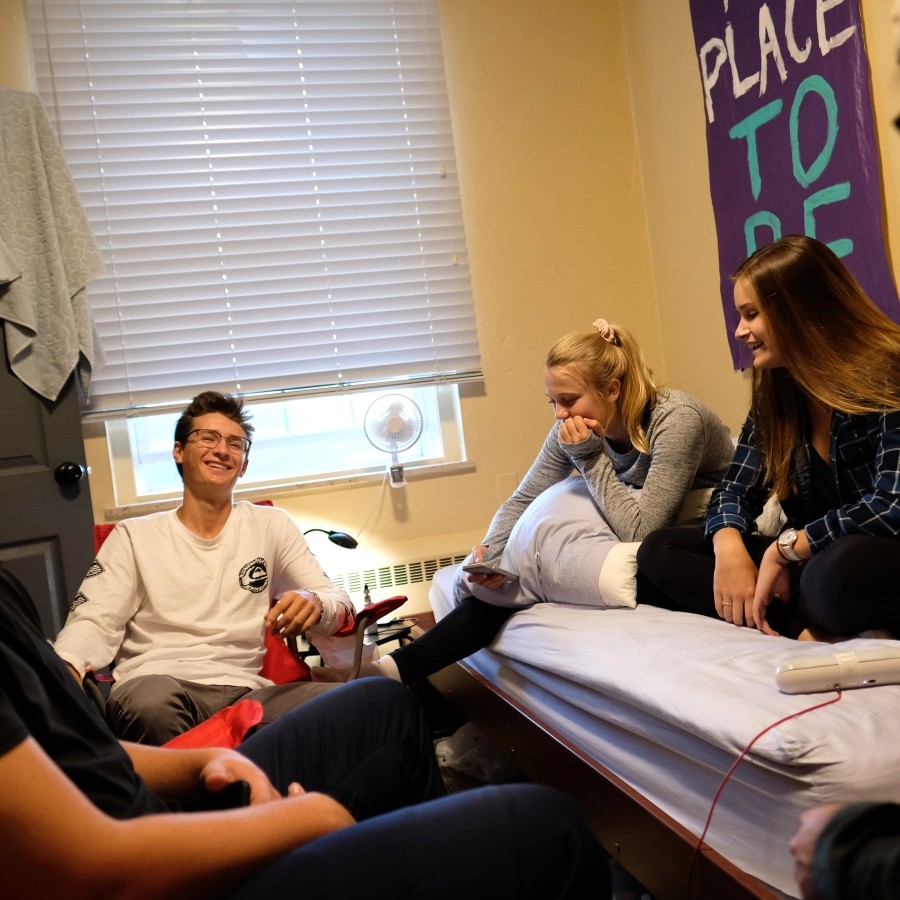Students socializing in a residence room. Two students are sitting on the bed, while the others are sitting on chairs.