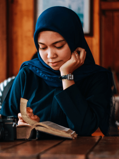 Woman in navy blue hijab reading a book at a wooden table